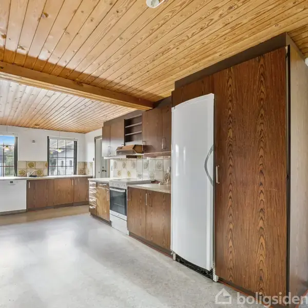 Køkken med træloft og lyse gulve. Mørke træskabe huser en indbygget ovn og køleskab. Vinduer giver naturligt lys, mens en murstensvæg til venstre tilføjer tekstur.