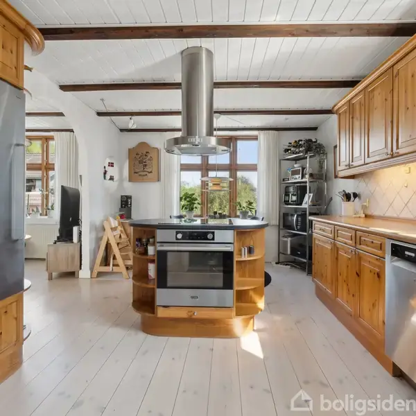 Køkken med kogeø i træ og stål, omgivet af skabe og hvide lofter med synlige bjælker. Store vinduer i baggrunden giver naturligt lys. Opvaskemaskine og diverse køkkenapparater på bordpladen.