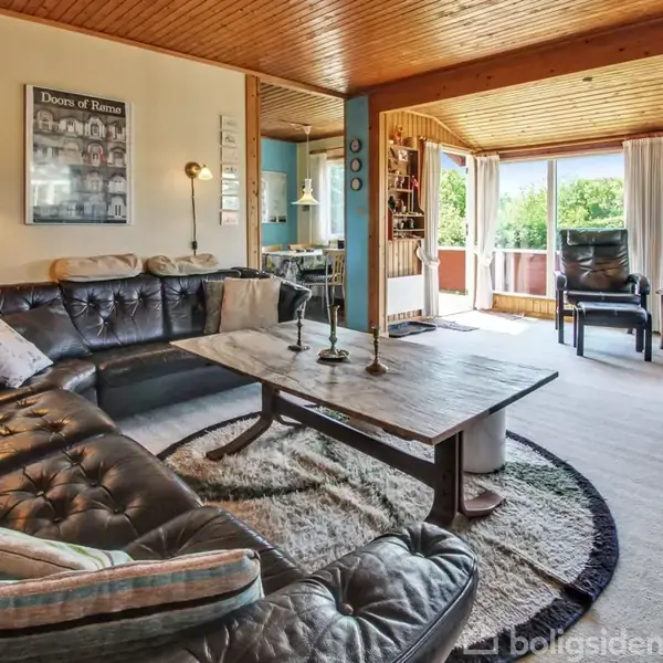 En sort lædersofa står i en stue med træloft. Sofaen omkranser et stort træbord. Rummet har store vinduer med udsigt til en have. En plakat med teksten "Doors of Rome" hænger på væggen.