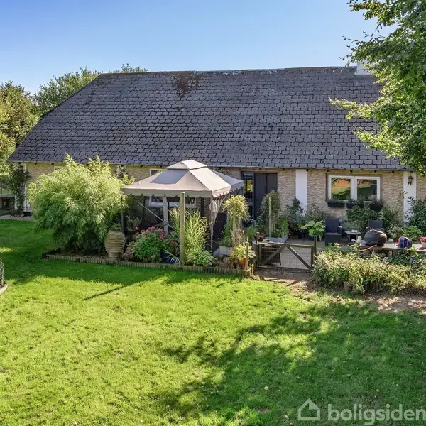 Hus med stenfacade og sort tag står i en frodig have. Foran huset er en terrasse med et hegn og en havepavillon. Omkringstående træer danner en grøn baggrund.