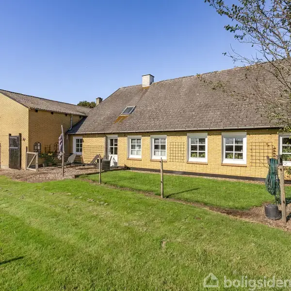 Gult bondehus med skrå tag står på en grøn græsplæne med blå himmel i baggrunden. Der er træer i højre side og en bygning til venstre.