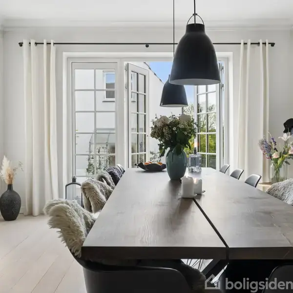Et spisebord i træ er dækket med blomster og stearinlys, omgivet af stole med pelspuder. To sorte lamper hænger over bordet i et lyst rum med store vinduer og et verdenskort på væggen.