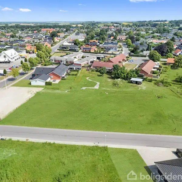 Åben, grøn mark ligger stille foran en vej, omgivet af spredte huse og et byområde i baggrunden. Spredte træer og forskellige bygninger danner en malerisk forstadsscene.