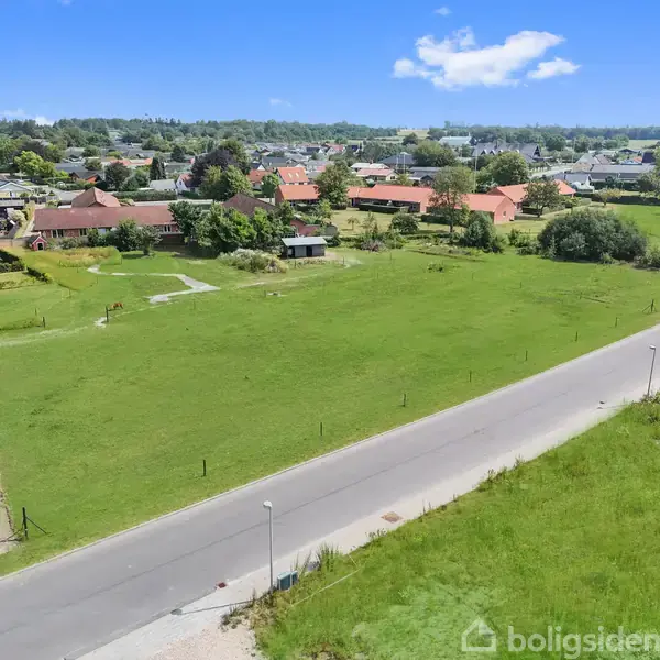Grønt græsareal ligger stille ved siden af en vej i et villakvarter med spredte huse og træer i baggrunden under en klar blå himmel.