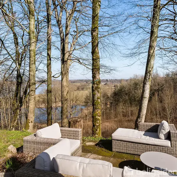 Udendørs sofaer er arrangeret omkring et lavt bord; de står på en terrasse med udsigt over en træbevokset eng og vandløb i klar solskinsvejr.