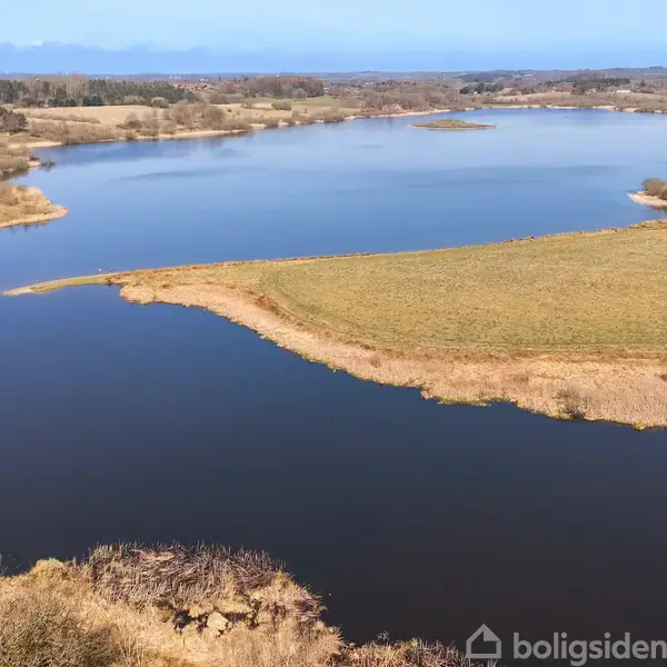 En stor sø strækker sig gennem landskabet, omgivet af åbne marker og skovområder under en klar blå himmel.