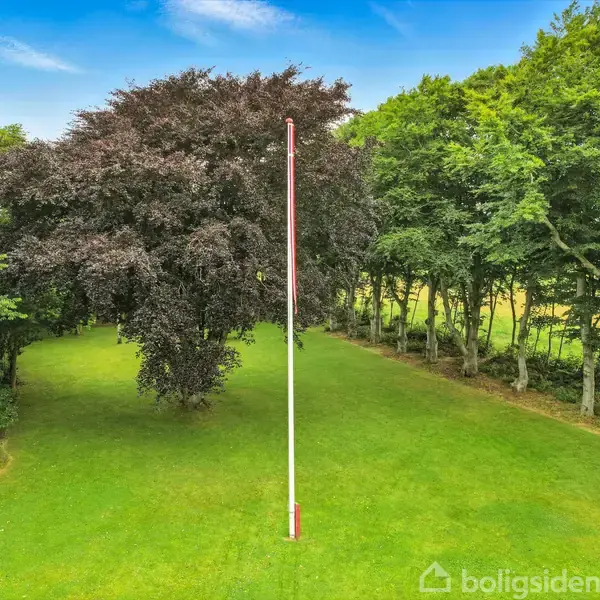 Flagstang står lodret på grøn plæne omgivet af træer og buske i en frodig park under en blå himmel med få skyer.