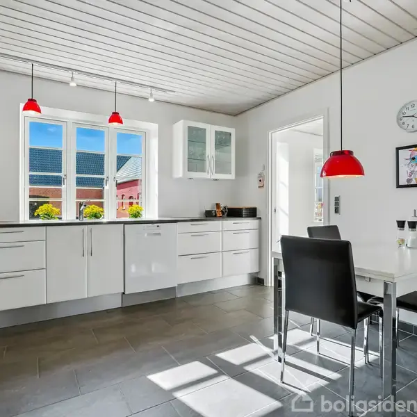 Køkken med hvide skabe og sort bordplade, har to røde pendellamper, et spisebord med fire stole, og et stort vindue, der slipper sollys ind fra en solrig dag.