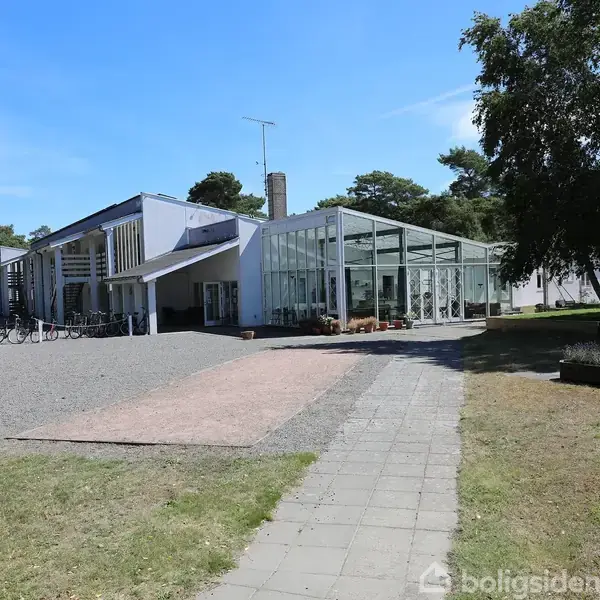 En moderne bygning står stille omgivet af grønne træer og græs. En cykel står udenfor, og en sti leder til en indgang ved siden af en glasudbygning.