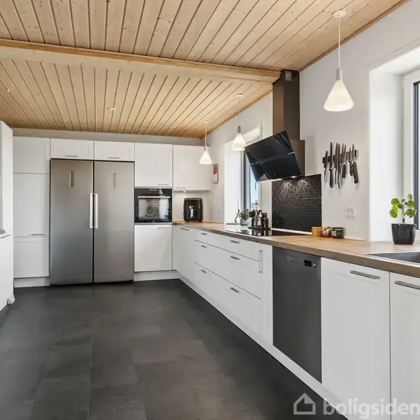 Køkken med hvide skabe og stål køleskab. To hængelamper lyser rummet op. Store vinduer giver udsigt til naturen udenfor. Skærebrætter og planter på bordplade ved vask.