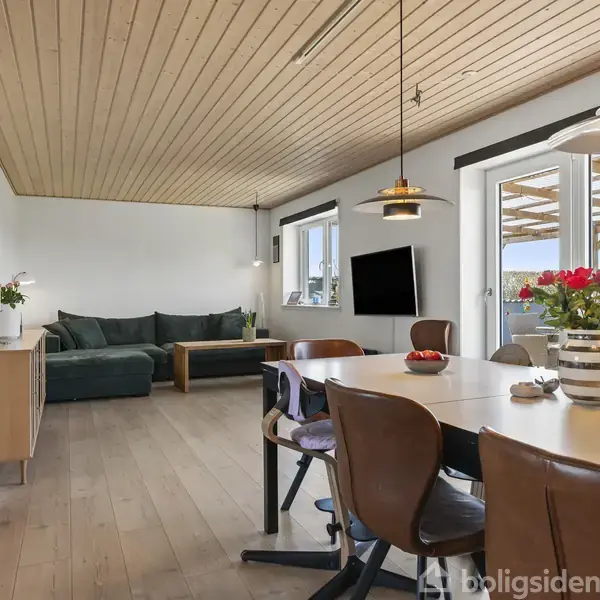 Stue med spisebord og stole, hvor en vase med røde blomster står centralt. I baggrunden ses en grøn sofa og et væghængt TV. Loftet og gulvet er i træ.