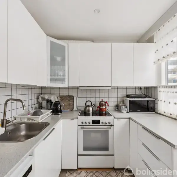 Et køkken med hvide skabe og fliser, hvor en komfur og en vask er placeret blandt kaffemaskiner og en mikrobølgeovn. Vinduesgardiner slipper lys ind fra udenfor.