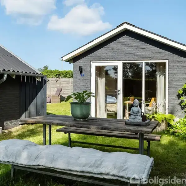 Et lille, mørkt gråt hus med glasdøre står i en have. Foran er et bord med en grøn plante og en Buddha-figur. Omkring er grønt græs og en træhegn.