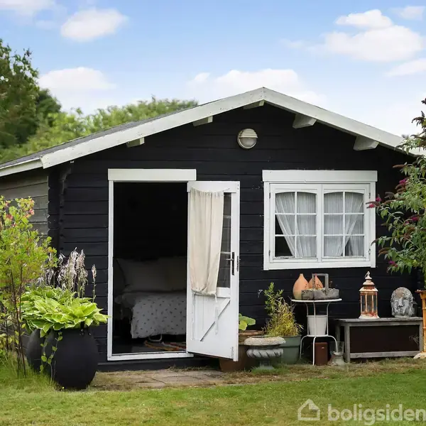 En sort træhytte står med åben dør, omgivet af frodig have med buske og blomster. Indenfor ses en seng med en hvid dyne. Sollys belyser scenen.