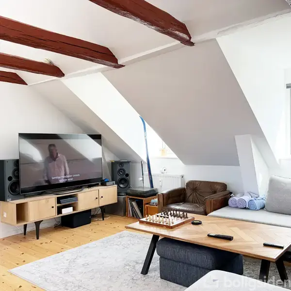 Et moderne stueområde med en fladskærms-tv, der viser tekst. Et sofabord med et skakbræt står foran en grå sofa. Træbjælker og skrå vægge skaber en hyggelig atmosfære.