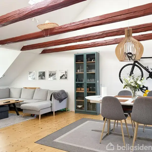 Gråt sofaarrangement står i en stue med træbjælker. Et spisebord med stole ses ved siden af en sort cykel på væggen. Et vitrineskab står i hjørnet.