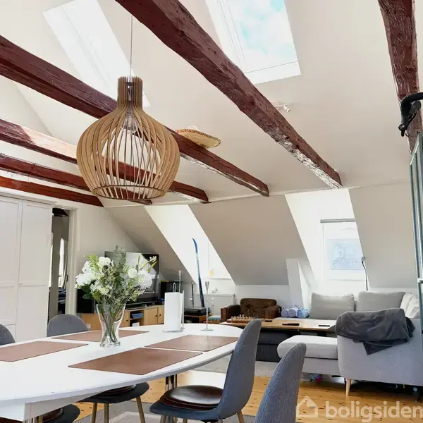 Et moderne spisebord med brune dækkeservietter er omgivet af stole i en stue under loft med bjælker. En lampe hænger over bordet, og sofaer findes i baggrunden.