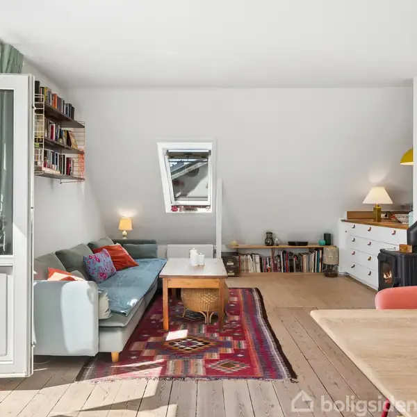 Stue med en blå sofa fyldt med farverige puder. Sofaen står på et tæppe med mønster. Bord og pejs i nærheden. Lyse vægge og trægulv. Hylde med bøger over sofa.