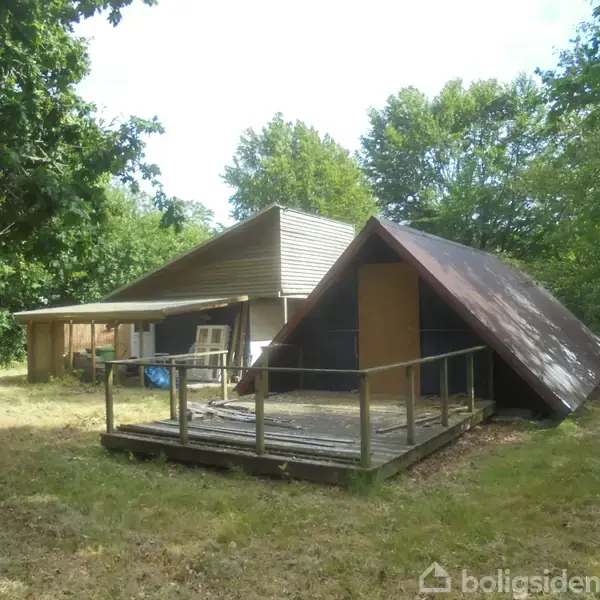 A-formet træstruktur står i græsbevokset have, omgivet af træer. Bygningen har terrasse med rækværk. Et større, fladt tag strækker sig ud bagved. Ingen tekst er synlig.