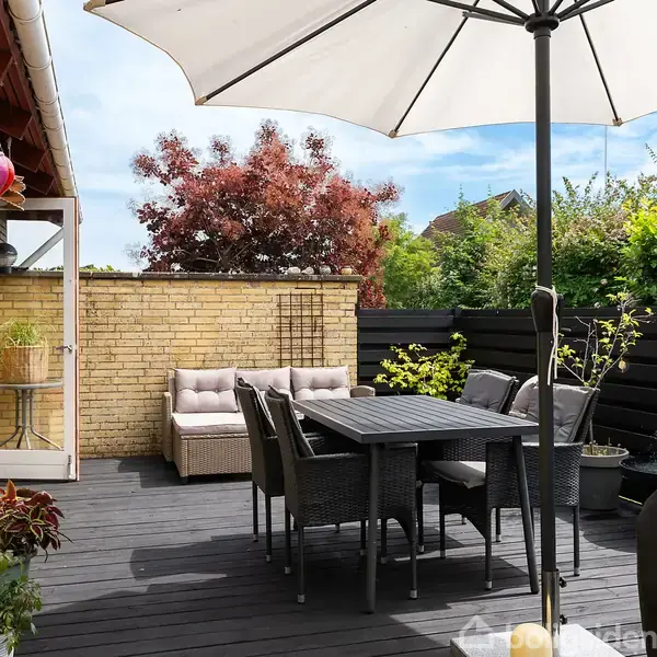 En udendørs terrasse med et mørkt trædæk har et bord og stole under en stor hvid parasol. Der er en sofa i hjørnet nær en murstensvæg, omgivet af grønt løv og buske.