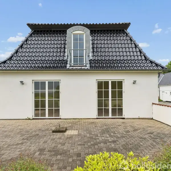 Et hvidt hus med sort tag og frontalt to store vinduer. Foran er en flisebelagt terrasse. Omgivet af grønne buske og træer i et åbent og klart vejr.