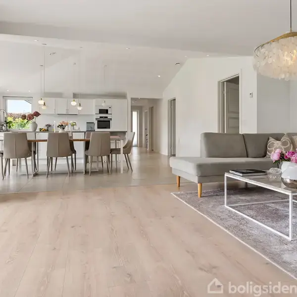 Stue med gråt hjørnesofa, sofabord med blomster, hængelampe og tæppe. Køkken med spisebord, stole og køkkenø i baggrunden. Lyse trægulve gennem hele rummet.