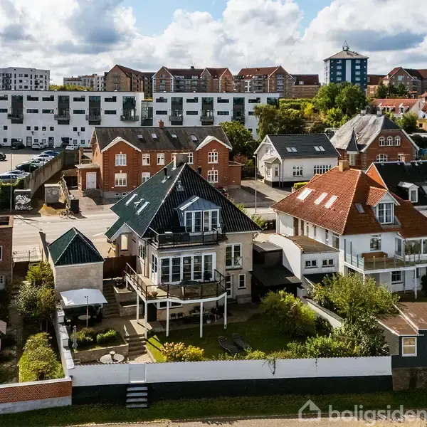 Et hvidt, to-etagers hus med sort tag står i et boligområde omgivet af lignende huse og grønne haver. Baggrunden viser flere røde murstensbygninger og moderne lejlighedskomplekser.