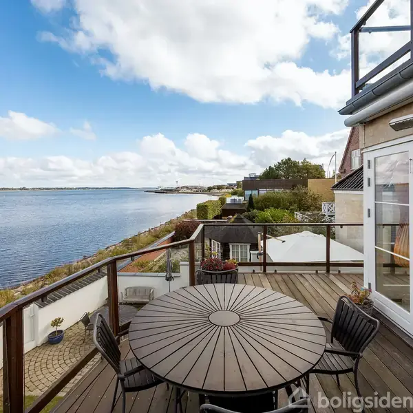 Rundt bord med seks stole står på en terrasse ved åbne franske døre, med udsigt over en bred flod eller fjord. Skyfri himmel og landskab dominerer baggrunden.