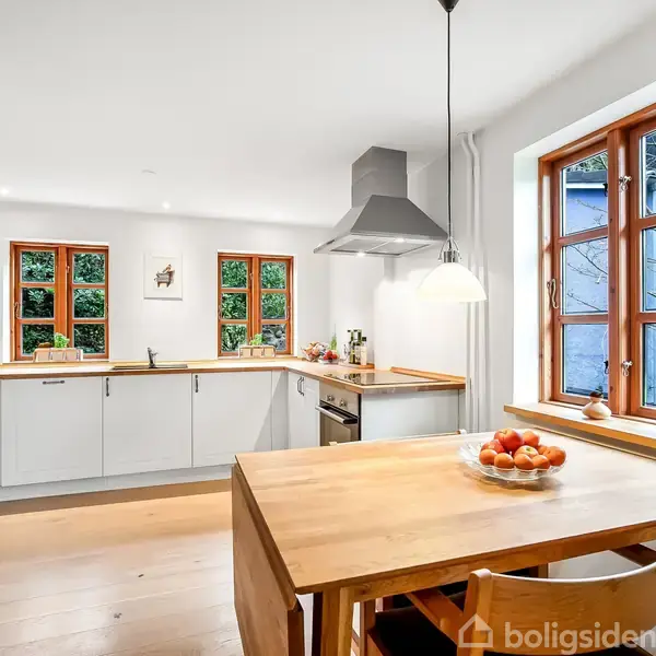 Et lyst køkken med hvide skabe, et træbord med en skål appelsiner, hængende lampe og store vinduer, der lukker naturligt lys ind. Murstensvæg til venstre skaber en rustik atmosfære.