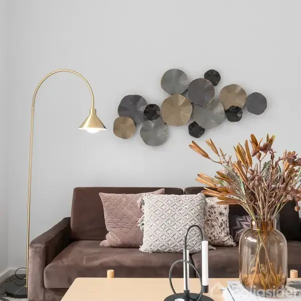 Brun sofa med lyse puder står foran en vægdekoration af cirkulære metalplader. En gulvlampe lyser rummet op, mens en vase med tørrede blomster står på sofabordet.