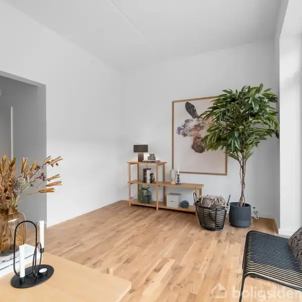 Et rummeligt værelse har en plante i en krukke ved siden af en reol med dekorationer. En sofa med puder står ved store vinduer. Et bord med vase og bøger er i forgrunden.