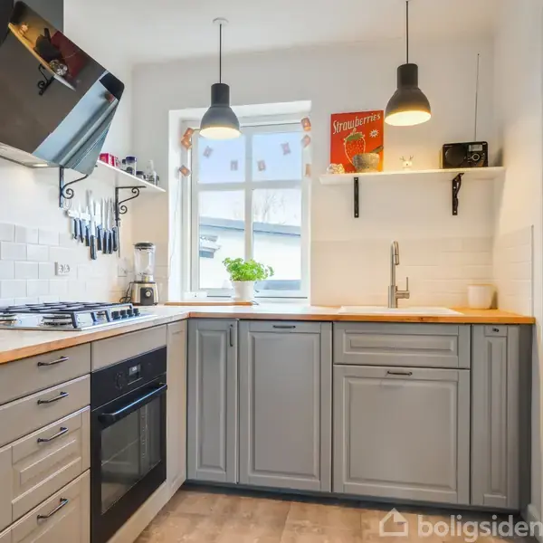 Grå køkkenskabe med træbordplade omringer en kogeplade. Et vindue med urtepotteskjuler i baggrunden. Over bordet hænger to lamper, og der står en "Stryhns"-pakke på hylden.