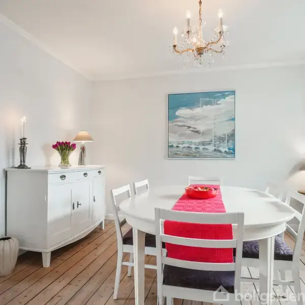 Spisebord med hvide stole står centralt, dekoreret med en rød løber. I baggrunden er et skab med blomster og stearinlys. Maleri hænger på væggen.