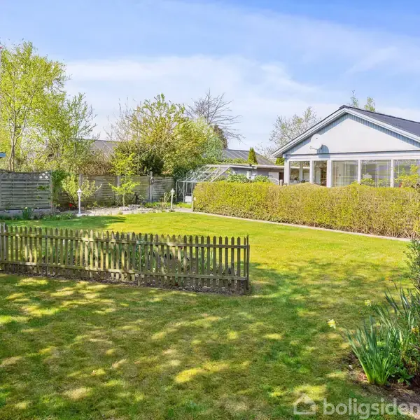 En velplejet have med et lavt hegn om et mindre område. En blå skurbygning står til venstre. En stor terrasse omgivet af hække er synlig foran huset.