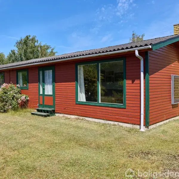 Et rødt træhus med grønne vinduesrammer og en gul murstensskorsten står på en velholdt græsplæne. Omkring huset er der spredt buskads og træer under en blå himmel.