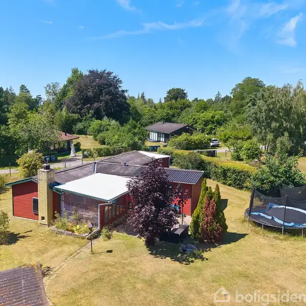 Et rødt træhus med fladt tag står i en grøn have. Ved siden af huset er der en trampolin på plænen omgivet af træer og buske under en blå himmel.