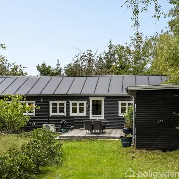 Sort sommerhus med hvidrammede vinduer står stille i en frodig have. Et træbord med stole er placeret på terrassen. Grønne buske og træer omgiver huset i naturlig idyl.