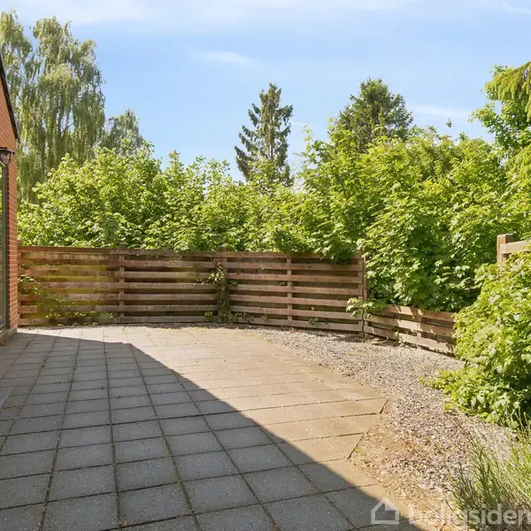 Terrasse med flisebelægning ved siden af en rød murstensbygning. Træhegn og tæt beplantning af buske og træer omkranser området, hvilket skaber en privat udendørs atmosfære.