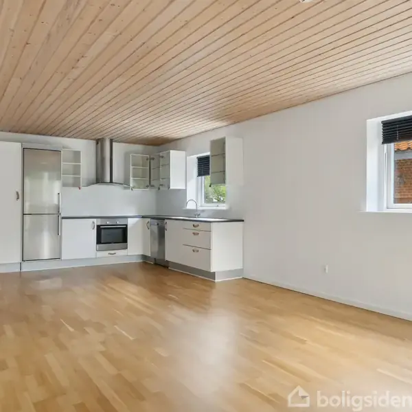 Køkken med hvide skabe og stålapparater, herunder et køleskab og ovn, placeret i en lys stue med trægulv og træloft. Enkelt vindue viser udsigt til en mursten bygning.