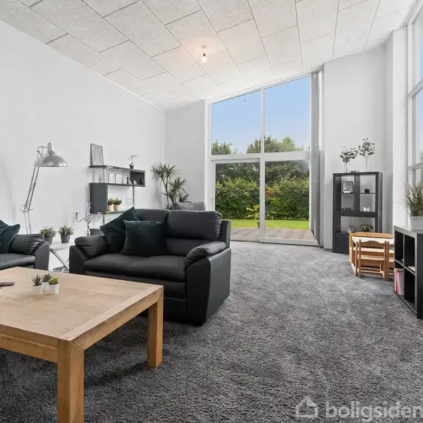 Sort lædersofa står i en stue med grå tæpper, et træsofabord og store vinduer, der giver udsyn til en grøn have. Reoler og planter pynter rummet.