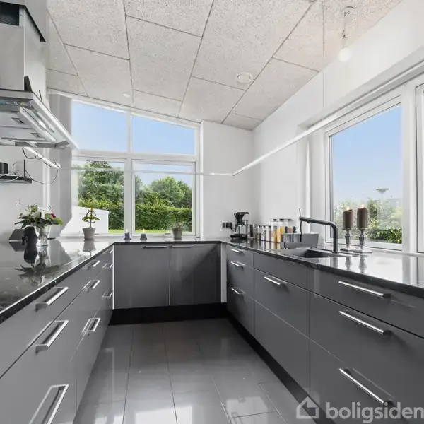 Et moderne køkken med L-formede grå skabe fyldt med køkkenredskaber, opdelt af en sort granitbordplade. Rum med store vinduer giver udsigt til grønne træer udenfor.