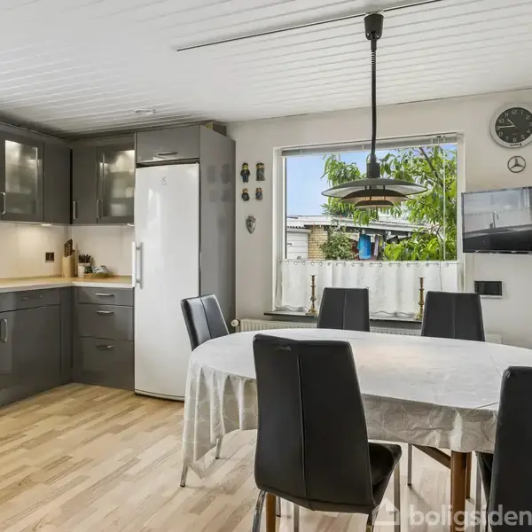 Et moderne køkken med grå skabe og hvidevarer, herunder et køleskab. En rund spisebord med sort stole står i midten. En lampe hænger over bordet, og et vindue viser en grøn udsigt.