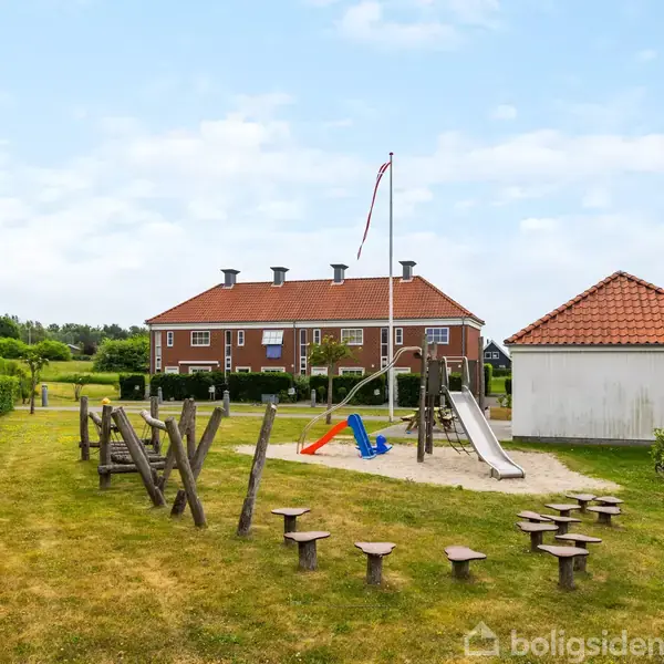 Legeplads med en rutsjebane og klatrestativer står på en sandet overflade. Omgivet af græsareal, rækker af huse og en hæk i baggrunden under en blå skyet himmel.