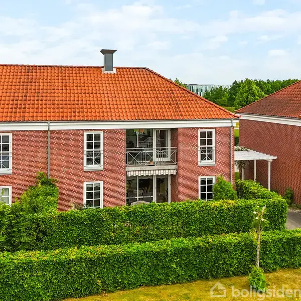 To røde murstenshuse med stejle, orange tegltage står stillet side om side. Husene omkranses af tætte hække og en grussti, med en blå himmel i baggrunden.