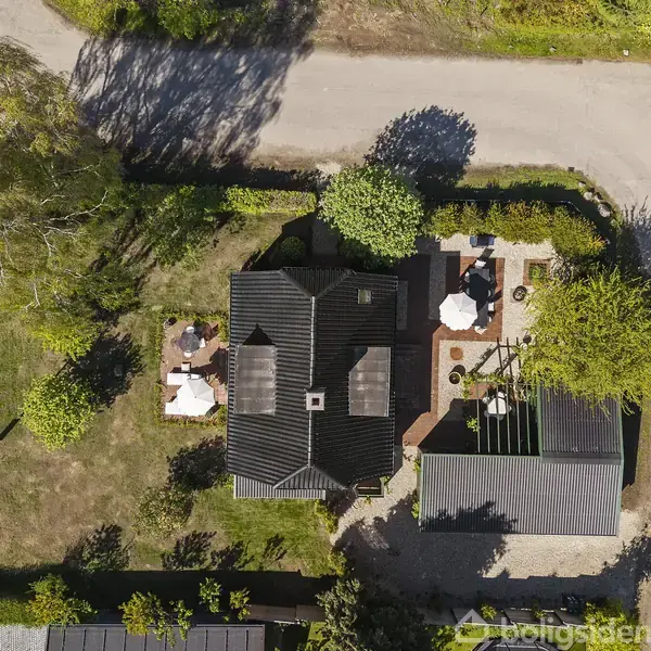 Hus set fra oven med mørkt tag omgivet af grønne træer og hæk. I haven er der terrasse med møbler og grusveje omkring.