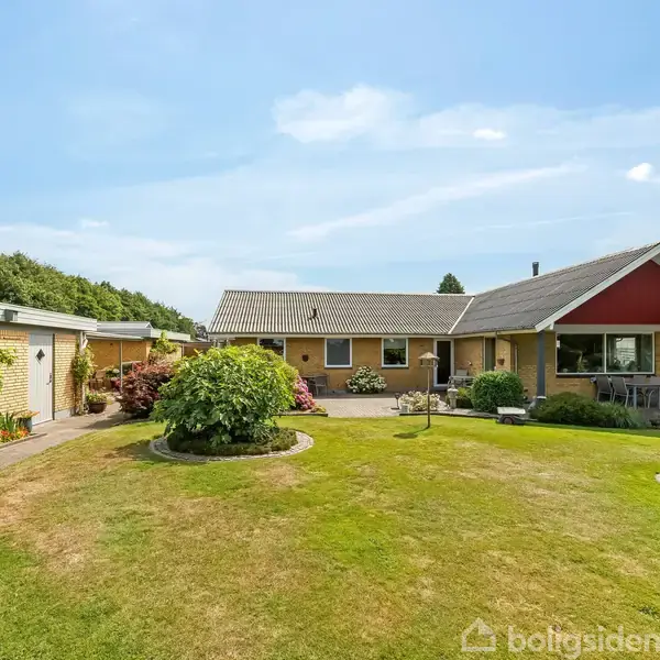 Et enfamiliehus ligger i en velholdt have med grøn plæne og farverige buske. Et flisebelagt område har havemøbler. Huset har gule murstensvægge og et skråt tag.