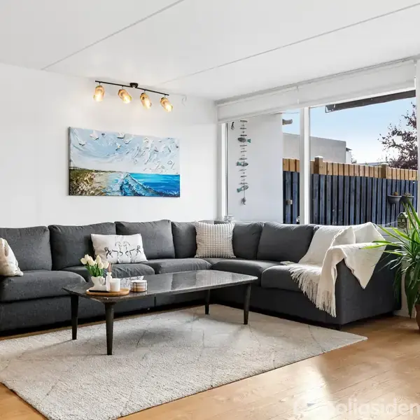 Stor, grå sofa med puder står i en moderne stue. Over sofaen hænger et maleri. Et sofabord står på et tæppe. Store vinduer slipper lys ind ved siden af en grøn plante.