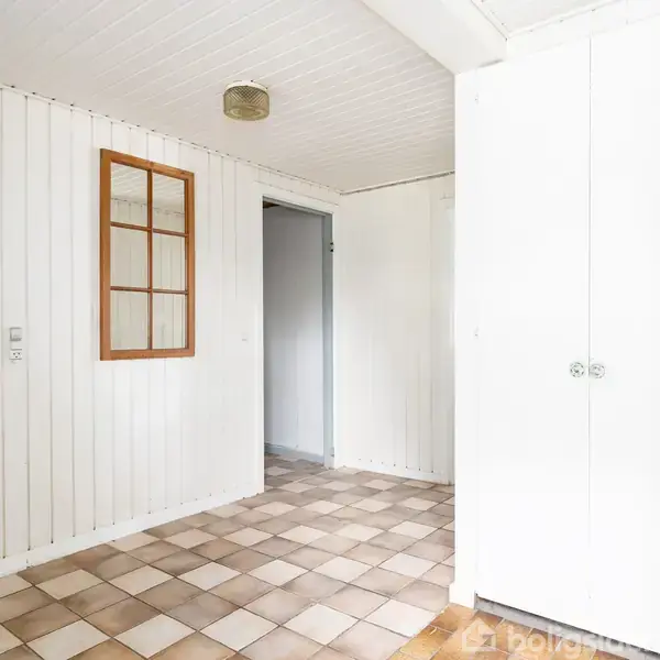 Et hvidt rum med flisegulv, skab på højre side, og døråbninger til venstre. Vindue i væggen fremhæver det enkle design. Badeværelse ses gennem døråbning.