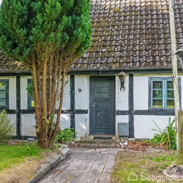 Bindingsværkshus med sort dør, omgivet af en have med græsplæne og buske. En lampepost står til højre for stien, der fører til hoveddøren. Husnummeret "1A" er ved døren.