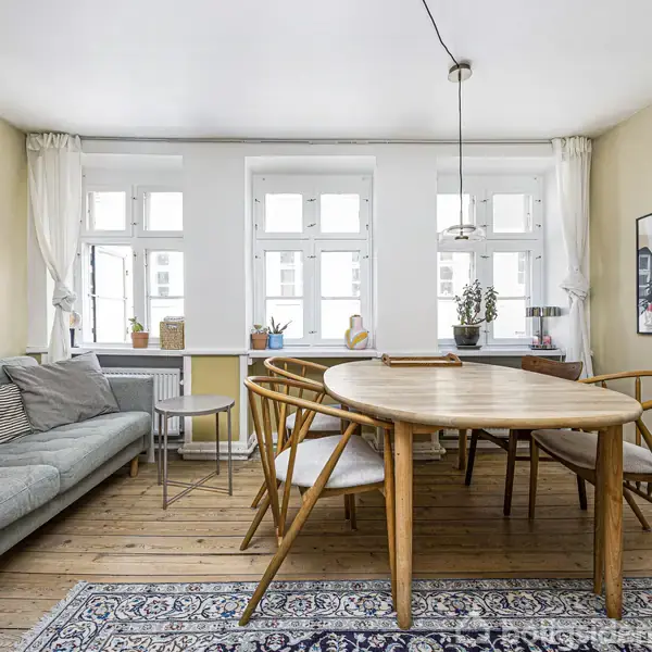 Sofagruppe i lysegråt står ved siden af et spisebord med stolper i træ. På væggen hænger plakaten "DAMASCUS". Store vinduer med hvidt gardin lukker lys ind i rummet.